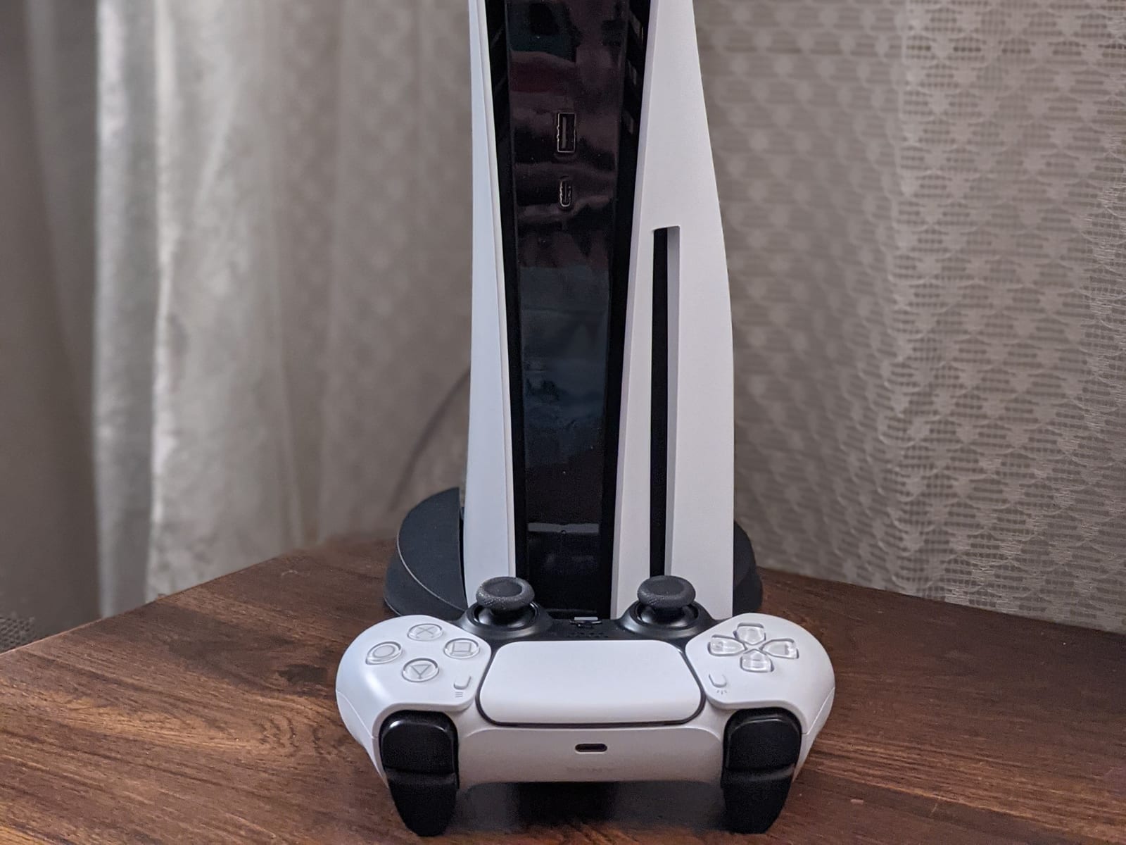 The lower half of a PlayStation 5 standing upright on its stand on a wooden table, with a white DualSense controller resting in front of it.