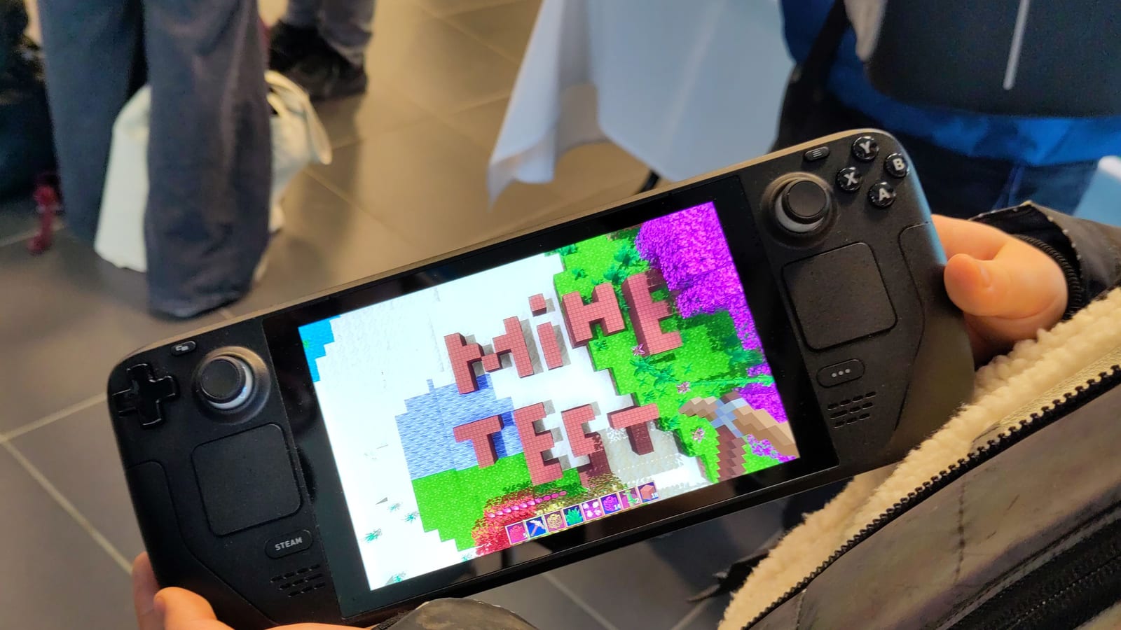 Two hands holding a Steam Deck handheld PC with the title screen of the game Minetest on its display, people standing in the background.