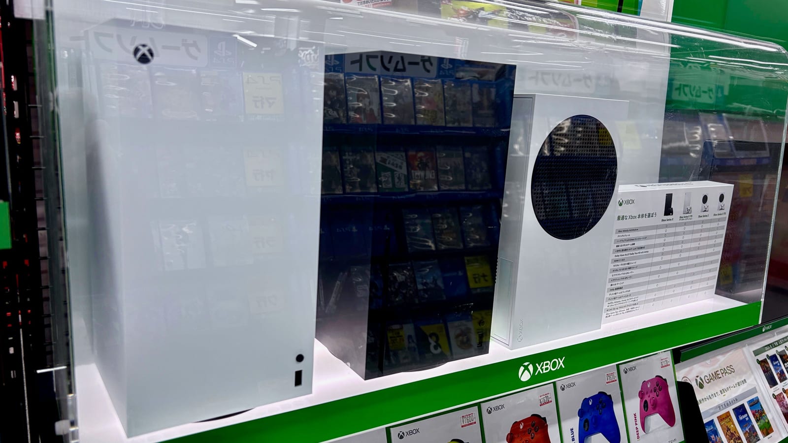 A white Xbox Series S and a larger Xbox Series X on a shop display shelf, with Xbox controllers and Game Pass cards on a green shelf below.